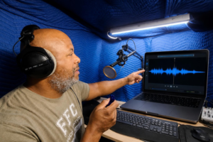 Cecil Archbold, voice actor, recording in his home studio booth with a laptop showing an audio waveform nearby